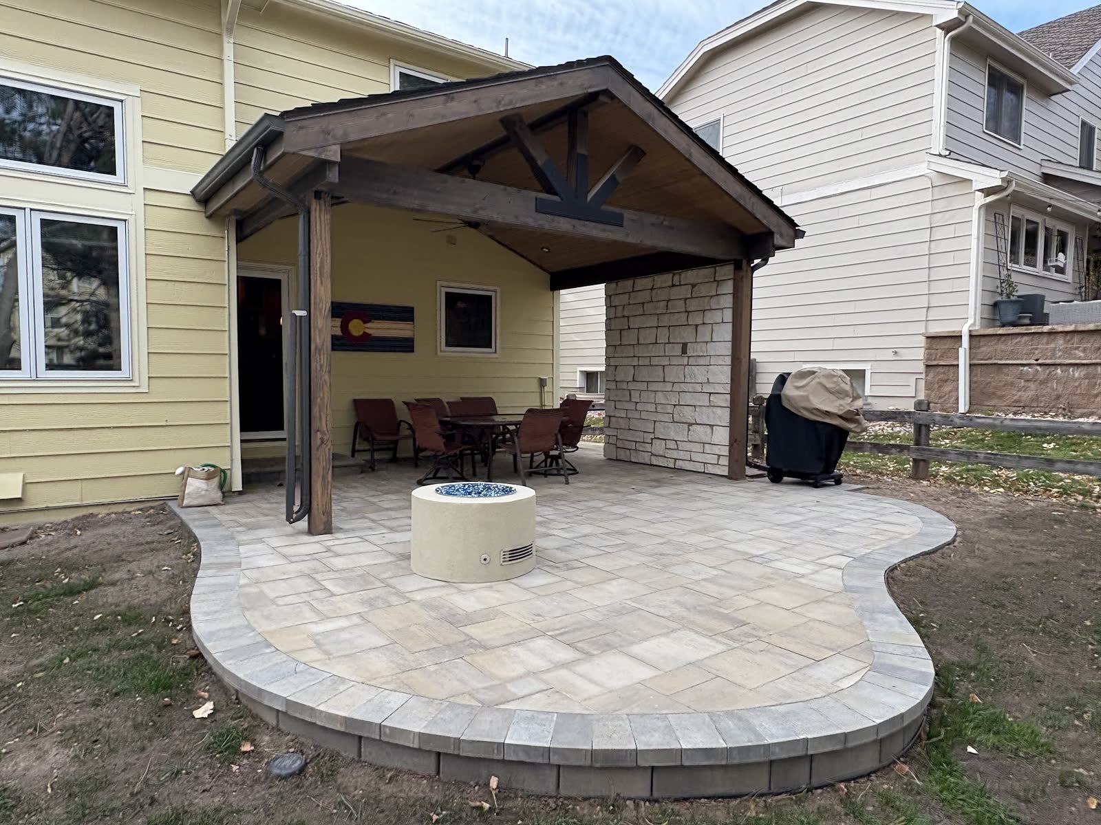 Paver patio with a round fire pit under a timber pavilion at dusk